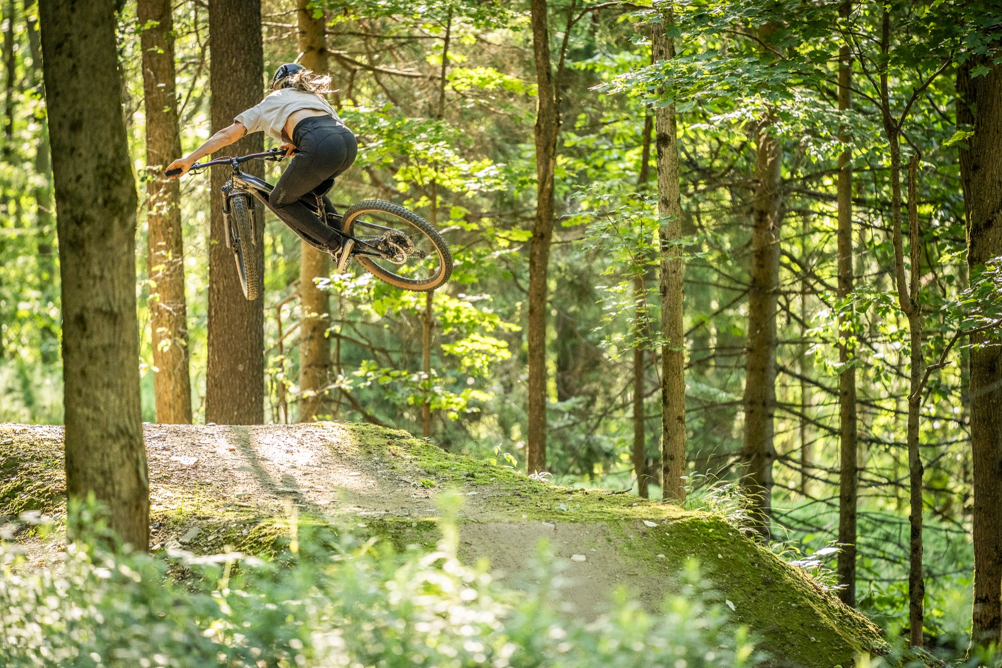 Mountain biker throwing a whip off a dirt jump in the sunlit summer woods at HoliMont in Ellicottville