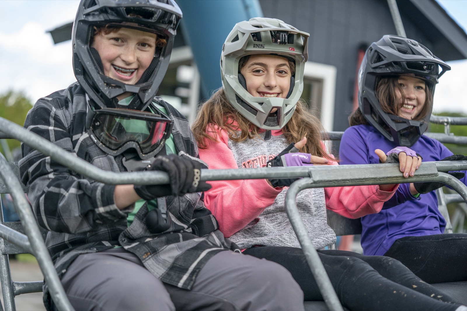 Three smiling kids in full-face helmets riding the chairlift at the HoliMont Bike Park